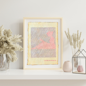 Handmade typewriter art on handmade, recycled, deckled-edged cotton paper. The repeated word “garden” is hand typed in black on a typewriter. The red text forms a visual of a watering can and small seedlings among the black text. The piece rests in a neutral frame on a white shelf styled with dried pampas grass, a glass terrarium, and pink textured vases to the sides. At the bottom of the art, the website “www.PlantsAreMyType.com” appears in red.