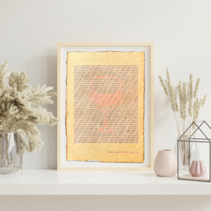 Handmade typewriter art on handmade, recycled, deckled-edged cotton paper. The repeated words “wine time” are hand typed in black on a typewriter. Red lettering forms a visual of a filled wine glass among the black text. The piece rests in a neutral frame on a white shelf styled with dried pampas grass, a glass terrarium, and pink textured vases to the sides. At the bottom of the art, the website “www.PlantsAreMyType.com” appears in red.