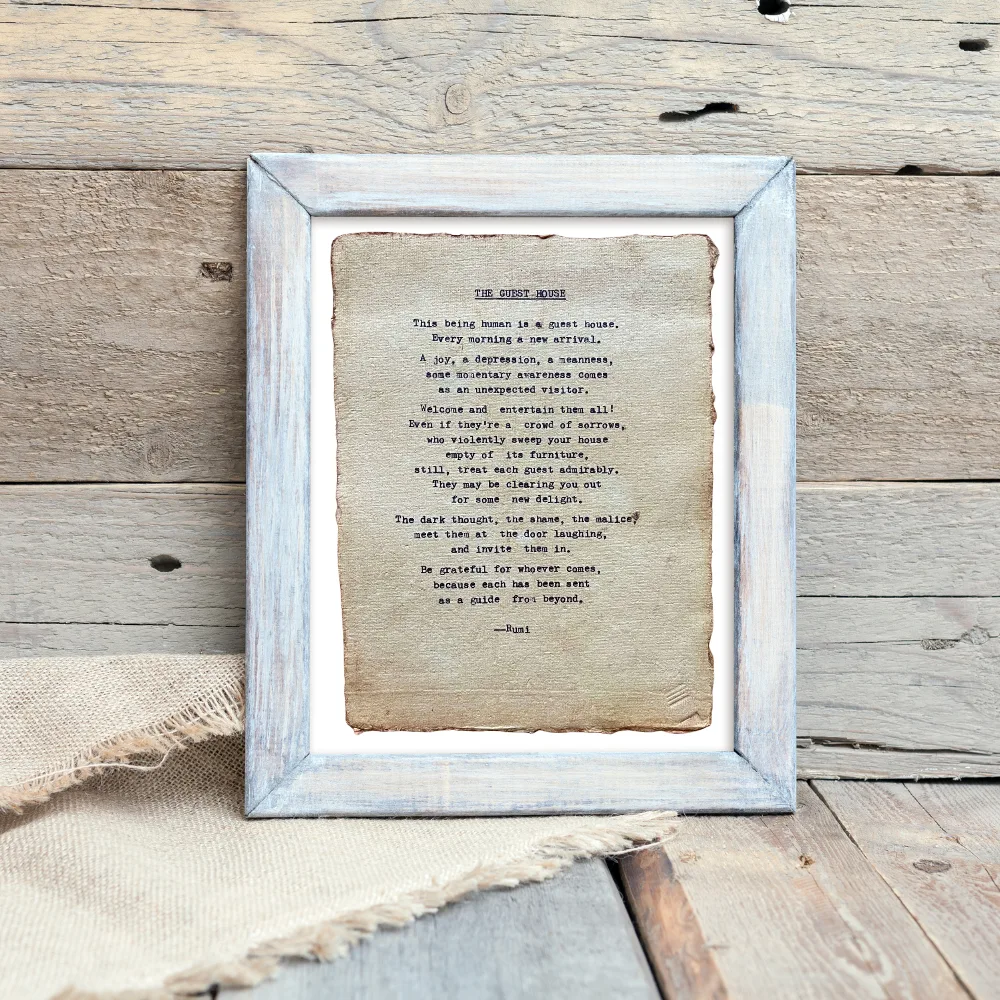 The guest house by Rumi poem hand typed on handmade, recycled cotton paper. The page is displayed in a blue wooden frame against a neutral, wooden background with a burlap fabric piece underneath.