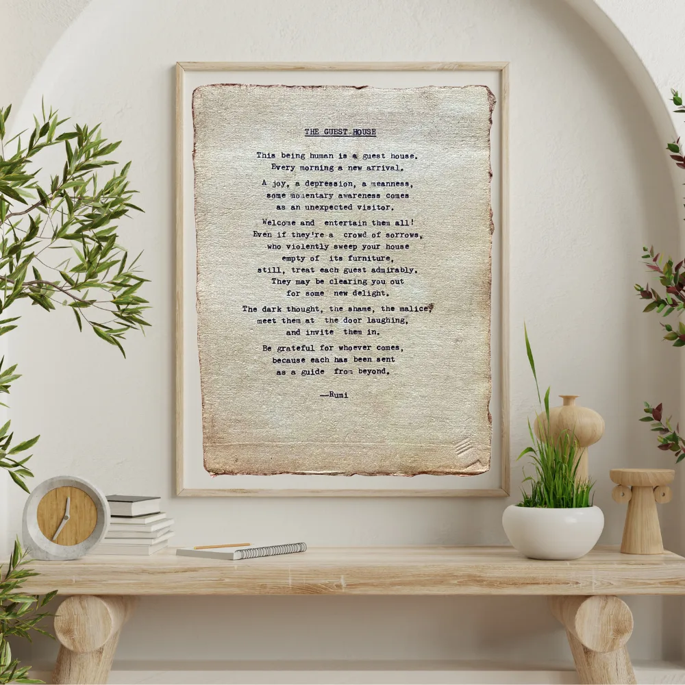 The guest house by Rumi poem hand typed on handmade, recycled cotton paper. The page is displayed in a neutral, wooden frame. The frame is above a wooden shelf displaying various greenery, a clock, books, and some wooden art.