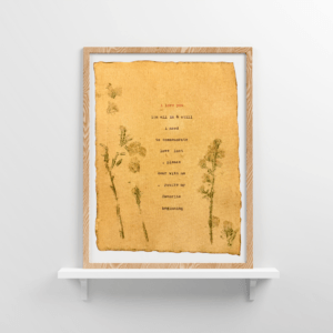 An image of framed typewriter art in a tan frame on a white floating shelf. The art is stamped with real flowers and displays a poem about love.