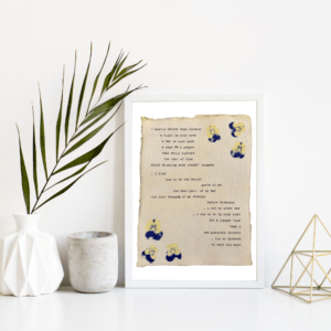 An image of framed typewriter art in a white frame on a white shelf. The art is stamped with real flowers and displays a poem about love. The art is displayed in a white frame between a white vase with a palm front next to a cement jar and a golden geometric decorative piece.