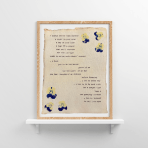 An image of framed typewriter art in a tan frame on a white floating shelf. The art is stamped with real flowers and displays a poem about love.
