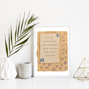 An image of framed typewriter art in a white frame on a white shelf. The art is stamped with real flowers and displays a poem about love. The art is displayed in a white frame between a white vase with a palm front next to a cement jar and a golden geometric decorative piece.
