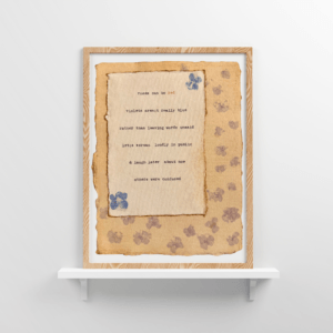 An image of framed typewriter art in a tan frame on a white floating shelf. The art is stamped with real flowers and displays a poem about love.