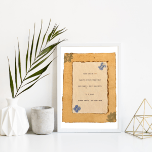 An image of framed typewriter art in a white frame on a white shelf. The art is stamped with real flowers and displays a poem about love. The art is displayed in a white frame between a white vase with a palm front next to a cement jar and a golden geometric decorative piece.