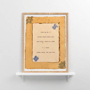 An image of framed typewriter art in a tan frame on a white floating shelf. The art is stamped with real flowers and displays a poem about love.