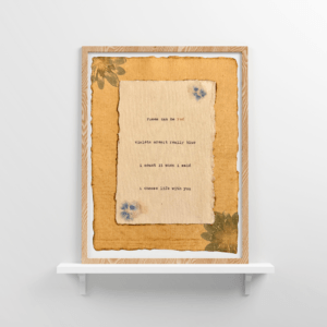 An image of framed typewriter art in a tan frame on a white floating shelf. The art is stamped with real flowers and displays a poem about love.