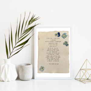 An image of framed typewriter art in a white frame on a white shelf. The art is stamped with real flowers and displays a poem about love. The art is displayed in a white frame between a white vase with a palm front next to a cement jar and a golden geometric decorative piece.
