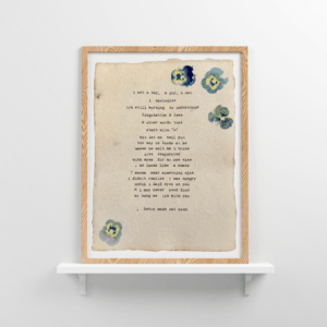 An image of framed typewriter art in a tan frame on a white floating shelf. The art is stamped with real flowers and displays a poem about love.