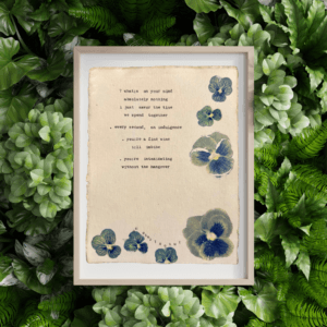 An image of framed typewriter art in a tan frame atop a background of greenery. The art is stamped with real flowers and displays a poem about love.