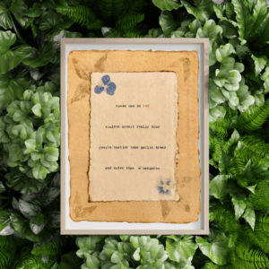 An image of framed typewriter art in a tan frame atop a background of greenery. The art is stamped with real flowers and displays a poem about love.