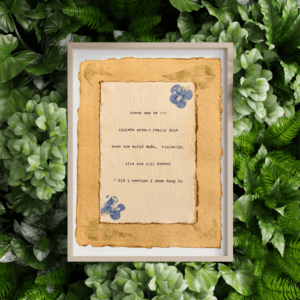 An image of framed typewriter art in a tan frame atop a background of greenery. The art is stamped with real flowers and displays a poem about love.