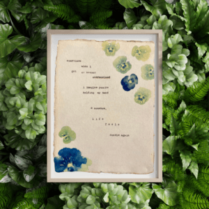 An image of framed typewriter art in a tan frame atop a background of greenery. The art is stamped with real flowers and displays a poem about love.