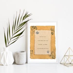 An image of framed typewriter art in a white frame on a white shelf. The art is stamped with real flowers and displays a poem about love. The art is displayed in a white frame between a white vase with a palm front next to a cement jar and a golden geometric decorative piece.