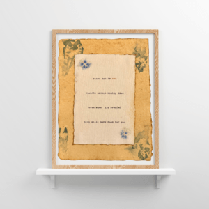 An image of framed typewriter art in a tan frame on a white floating shelf. The art is stamped with real flowers and displays a poem about love.