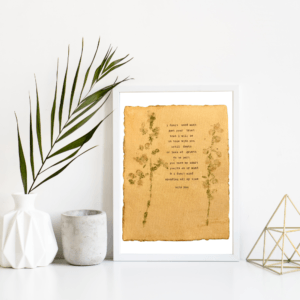 An image of framed typewriter art in a white frame on a white shelf. The art is stamped with real flowers and displays a poem about love. The art is displayed in a white frame between a white vase with a palm front next to a cement jar and a golden geometric decorative piece.