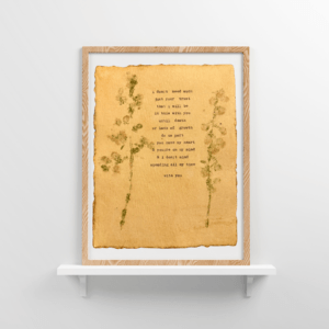 An image of framed typewriter art in a tan frame on a white floating shelf. The art is stamped with real flowers and displays a poem about love.