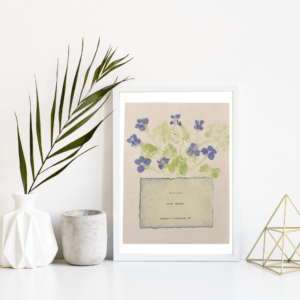 An image of framed typewriter art in a white frame on a white shelf. The art is stamped with real plants and displays a the plant name, botanical name, and area the plants were collected. The art is displayed in a white frame between a white vase with a palm front next to a cement jar and a golden geometric decorative piece.